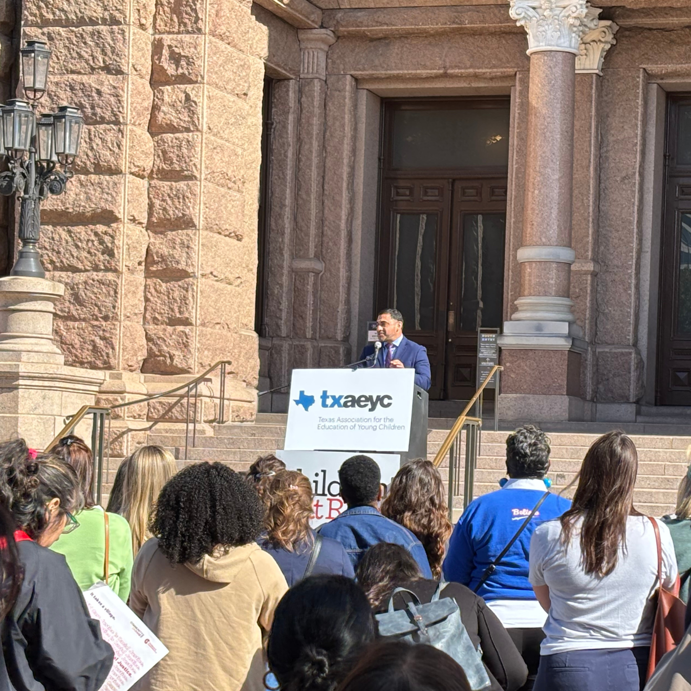 A speaker addresses a crowd gathered at the Texas State Capitol, with a podium displaying the Texas Association for the Education of Young Children logo. People in the audience are listening attentively.